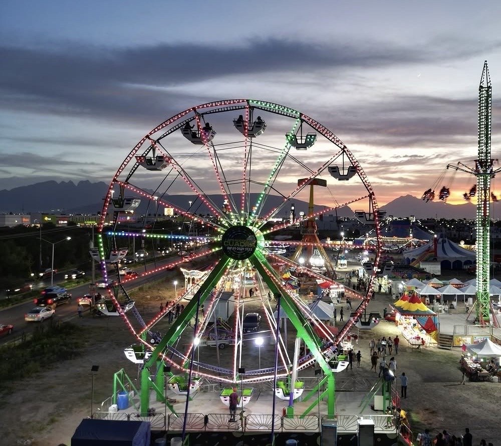 Vista aérea de una feria montada al atardecer: rueda de la fortuna con el logotipo de Cuadra, carpas blancas, puestos y la torre de sillas voladoras, con montañas en el horizonte